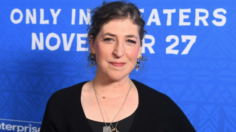 Wearing a black ensemble and different sets of jewelry, Mayim Bialik squints at the camera while smiling.