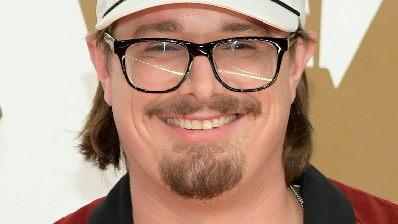 HARDY, smiling, on a red carpet, wearing glasses, wearing a white hat, facial hair