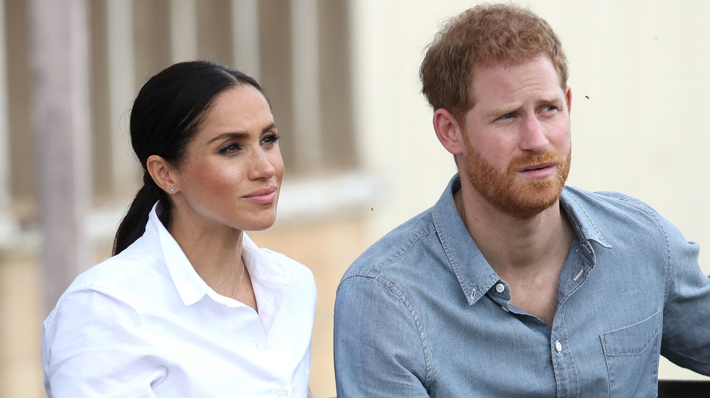 Meghan Markle in white sits next to Prince Harry in a jean shirt as they look out pensively at an event