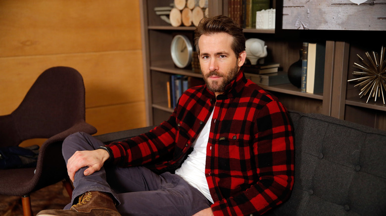 A bearded Ryan Reynolds sitting with his leg crossed, wearing plaid