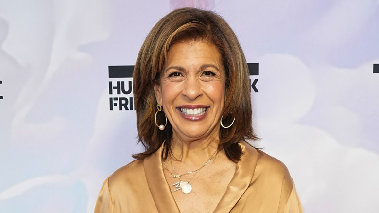 Hoda Kotb smiles while wearing a beige silk top, stacked necklaces, and hoop earrings.