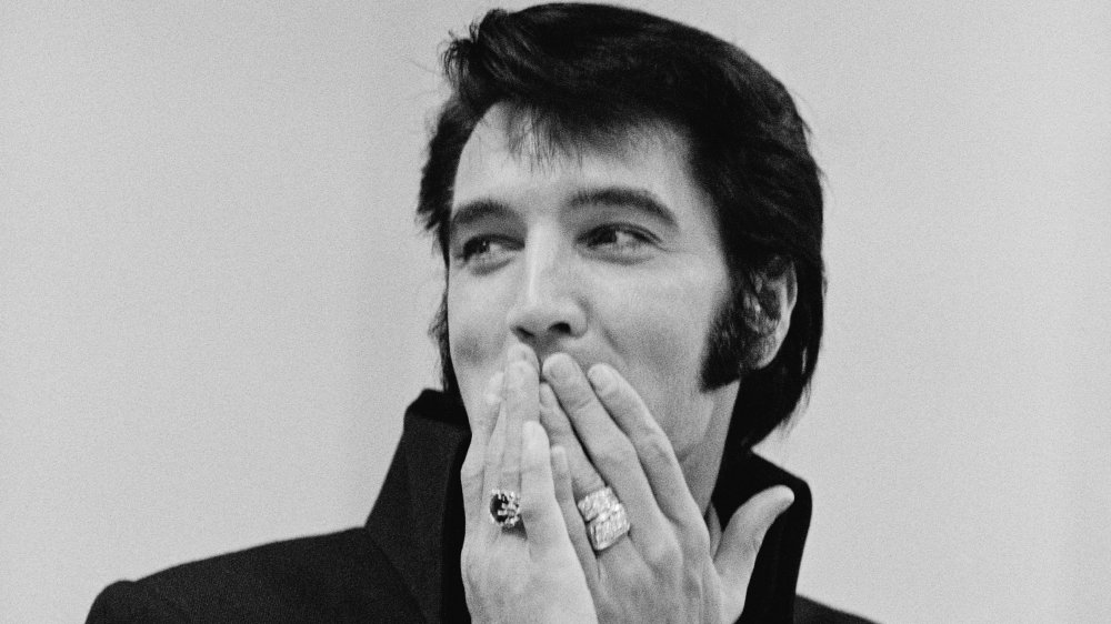 A black and white photo of Elvis Presley wearing rings and with sideburns covering his mouth with his hands