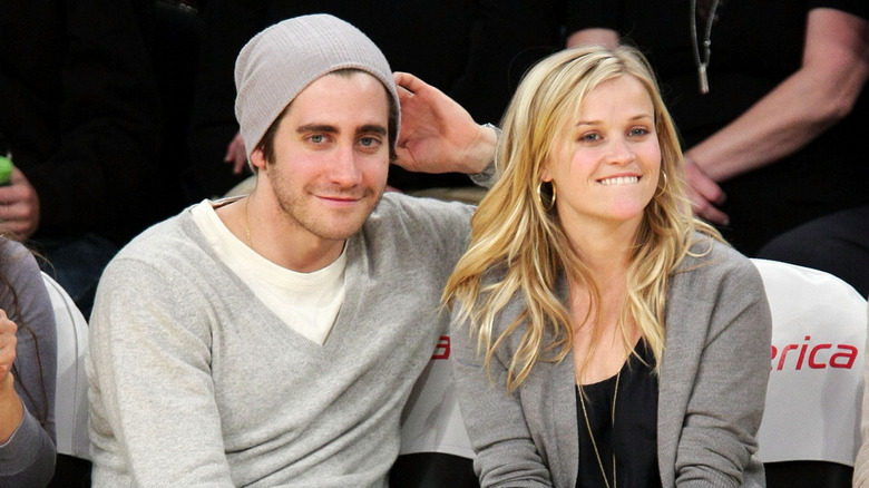 Jake Gyllenhaal wearing a grey long-sleeved shirt, white t-shirt, and grey beanie with Reese Witherspoon wearing a grey cardigan and black t-shirt
