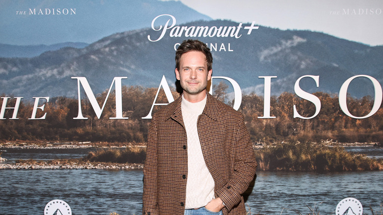 Patrick J. Adams posing against a backdrop for "The Madison" with one hand in the pocket of his jeans