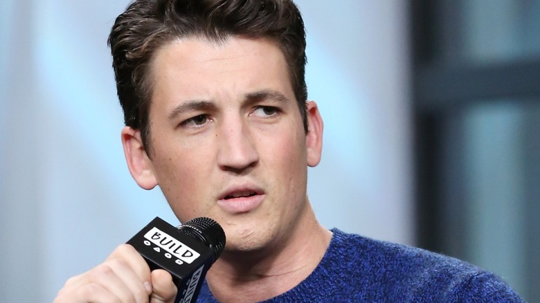 Miles Teller frowns while speaking in a microphone.