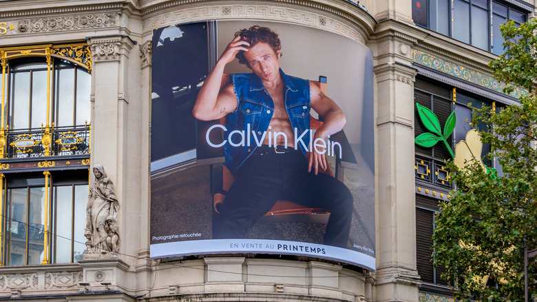 In a Calvin Klein ad on a billboard on the side of a building, Jeremy Allen White sits in a chair and poses