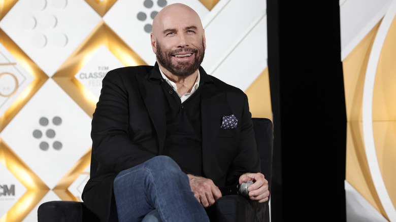 John Travolta smiling while sitting on stage in a black suit holding a microphone