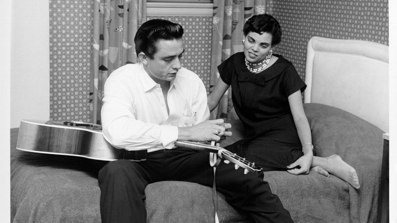 Johnny Cash holding a guitar sitting next to Vivian Liberto on a bed.