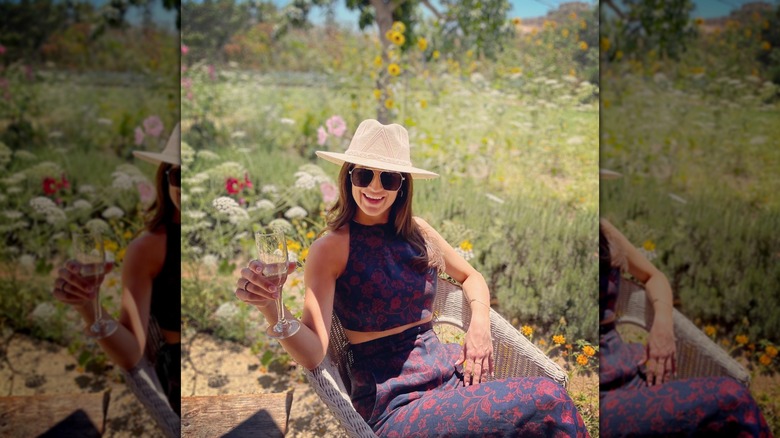 Kaitlan Collins sitting in chair with flowers behind her and holding a drink.
