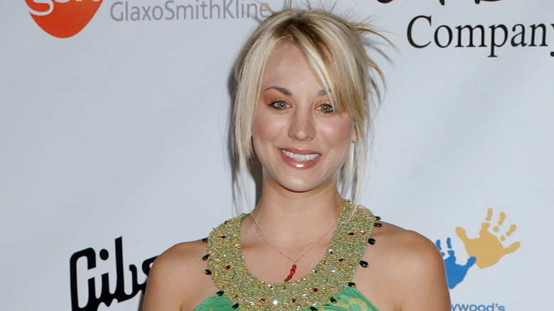 Kaley Cuoco wears a green bejeweled halter top, orange smoky eye shadow, and a blonde hair piece