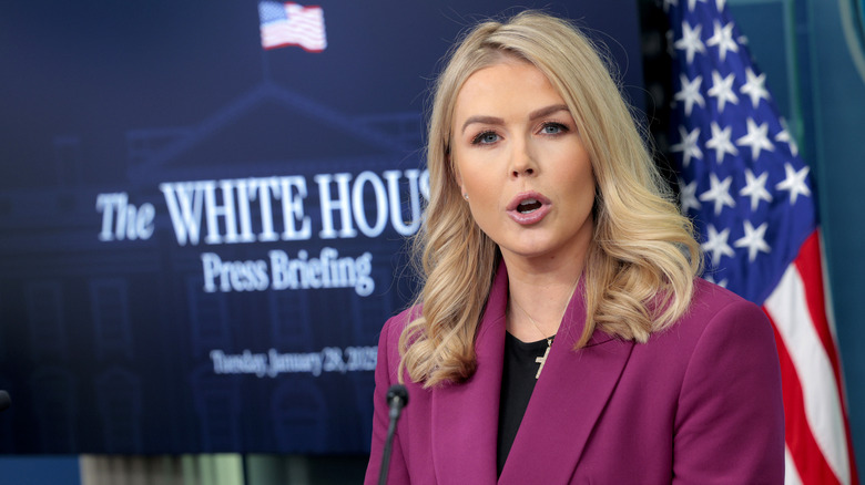 Karoline Leavitt speaking from the podium in the White House press briefing room in a plum blazer