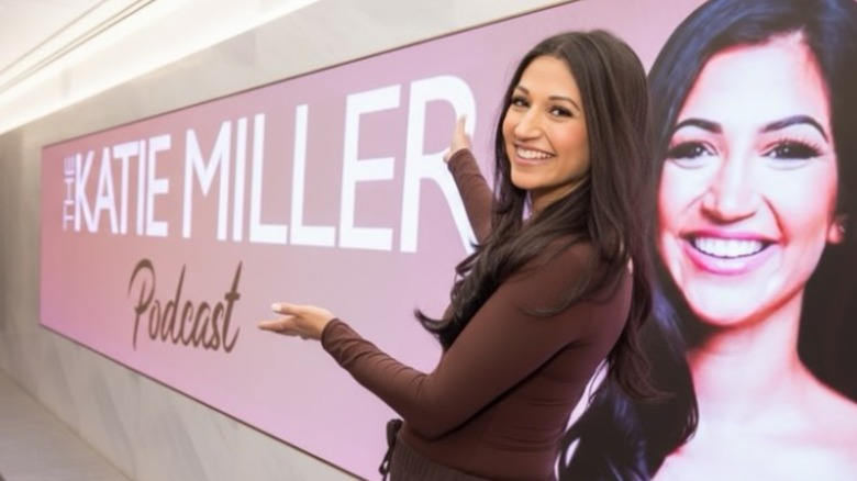 Katie Miller smiling while proudly posing with a banner for her self-titled podcast