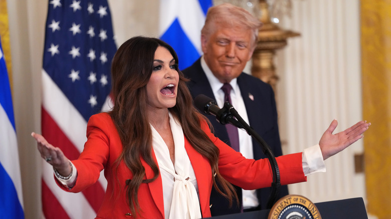 Kimberly Guilfoyle speaking next to Donald Trump in front of Greek and U.S. flags