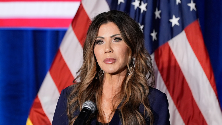Kristi Noem poker face in front of a microphone and an American flag