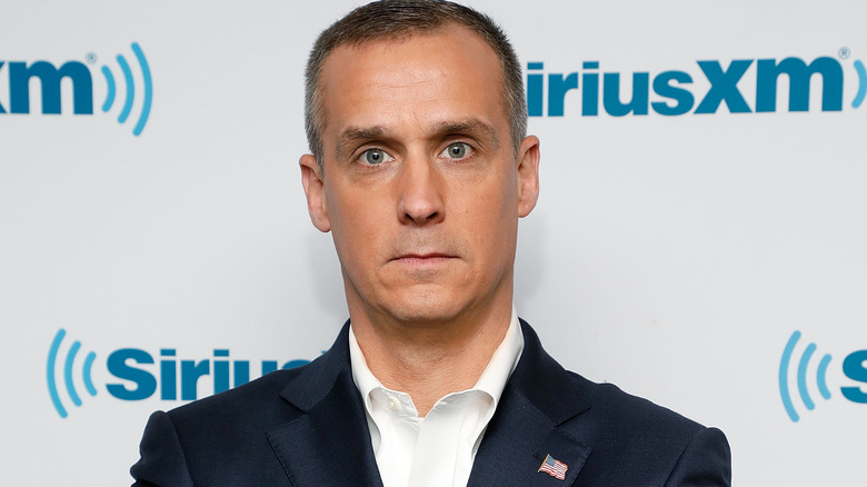 Corey Lewandowski looking gormless in a navy suit and an open-collared white shirt with an American flag pin