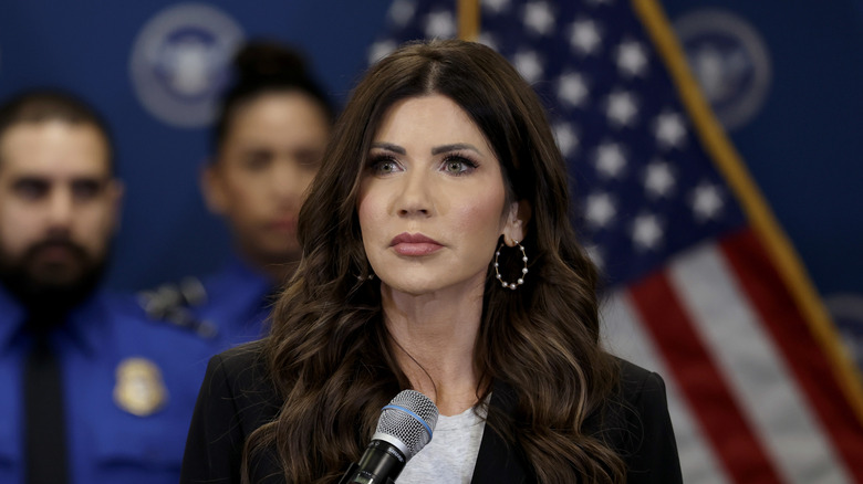 Kristi Noem listening during a press conference in front of a microphone in a black blazer