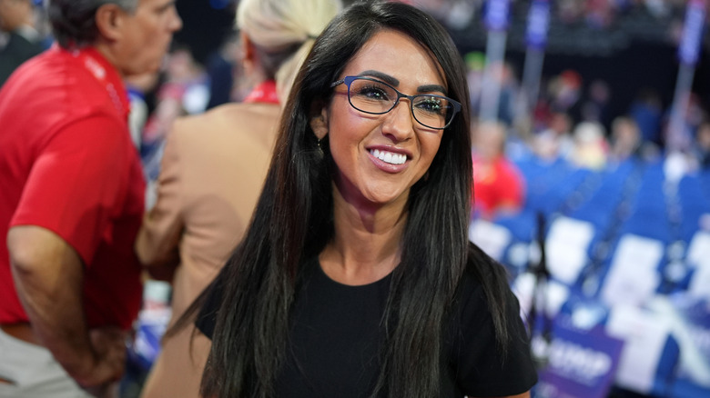 Lauren Boebert with arena seating behind her, wearing glasses, hair down, lots of makeup, looking to the side and smiling