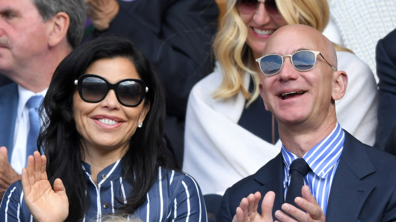 Lauren Sánchez and Jeff Bezos smiling and clapping while wearing sunglasses