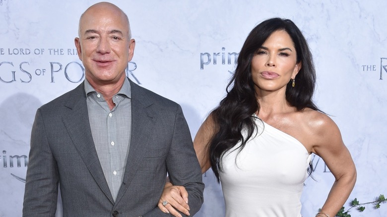 Jeff Bezos is all smiles as Lauren Sanchez, whose lips are pouted, wraps her arm around his.