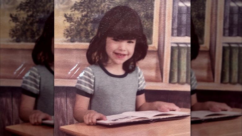 Lauren Sánchez as a child smiling and holding an open book on a desk in a classroom.