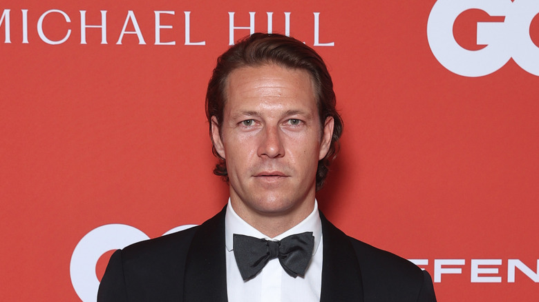 Luke Bracey staring straight into the camera in a tuxedo against a red background