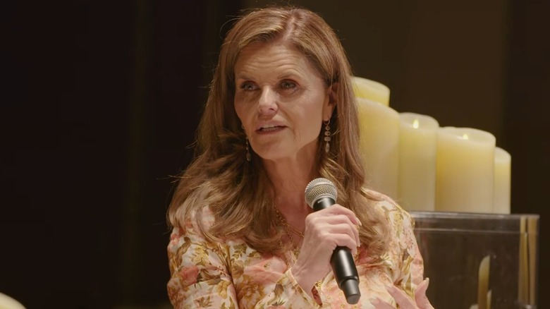 Wearing a flowery number and golden jewelry, Maria Shriver holds a microphone while speaking