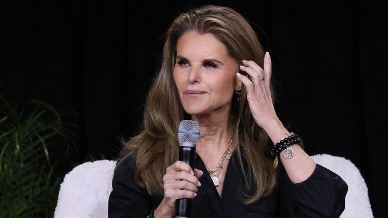 Maria Shriver holds a microphone with one hand and brushes her hair with the other