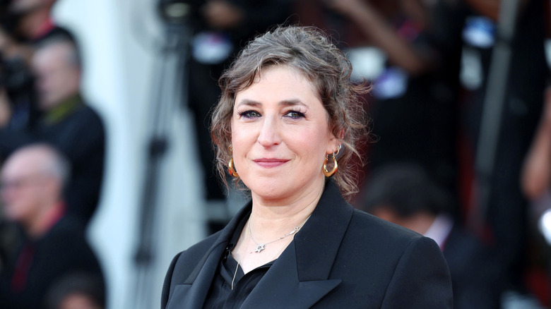 Mayim Bialik smiling in a black blazer with her hair pulled up.