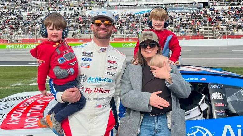Kelly LaJoie and Corey LaJoie smiling in sunglasses in front of Corey's car beside a racetrack