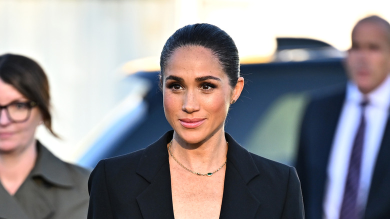 Meghan Markle smiling while wearing a black blazer with her hair in a tight, low bun