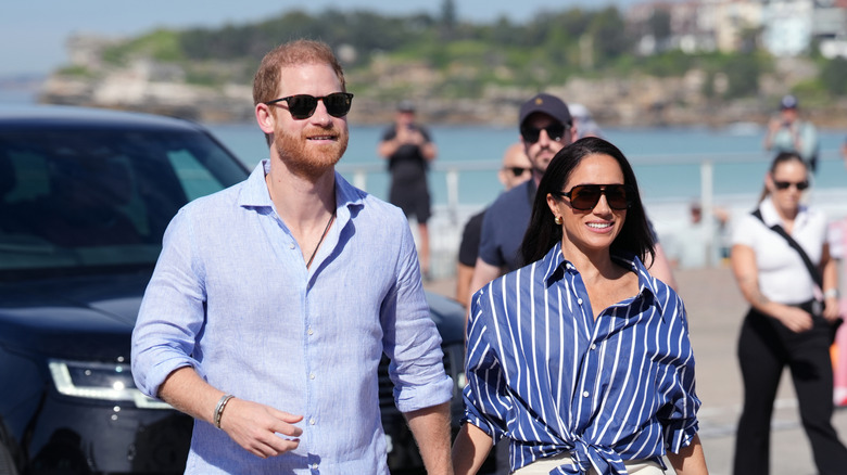 Prince Harry and Meghan Markle walking and smiling while holding hands in sunglasses and complementary outfits
