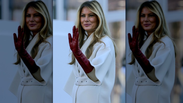 Melania Trump waving to a crowd in a white coat with red leather gloves