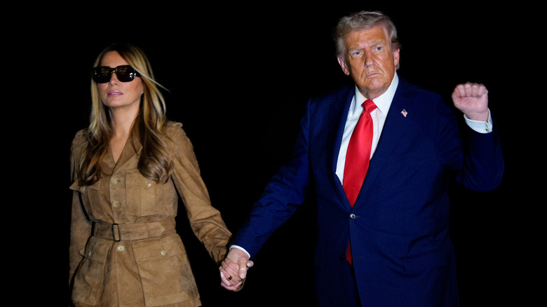 Donald Trump holding up a fist while walking next to and holding hands with Melania Trump in sunglasses