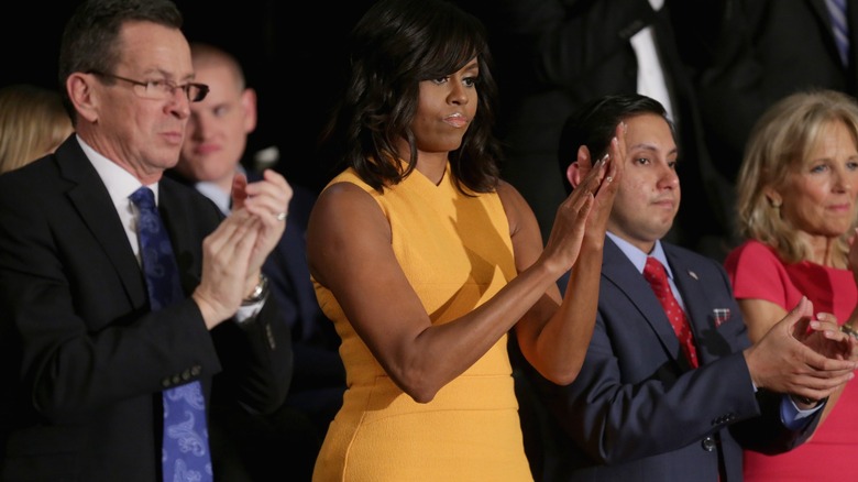 Wearing a sleeveless, fitting marigold dress, Michelle Obama claps alongside others while gazing downward.