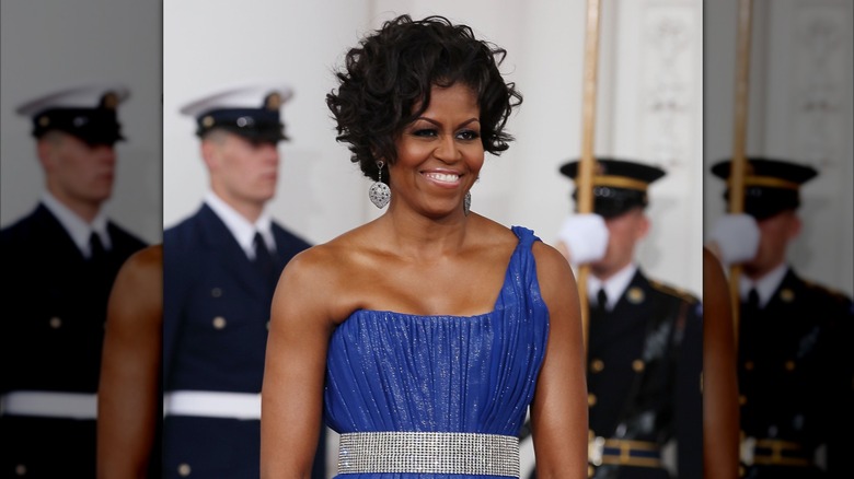 Wearing a pleated purple gown with a tight neckline, Michelle Obama smiles while looking into the distance.