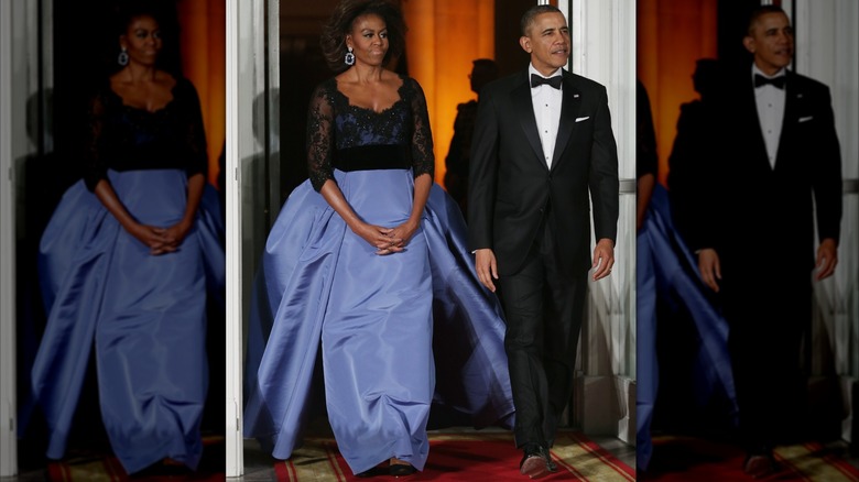 Wearing a black and purple ballgown, Michelle Obama walks next to Barack Obama with her hands clasped together.