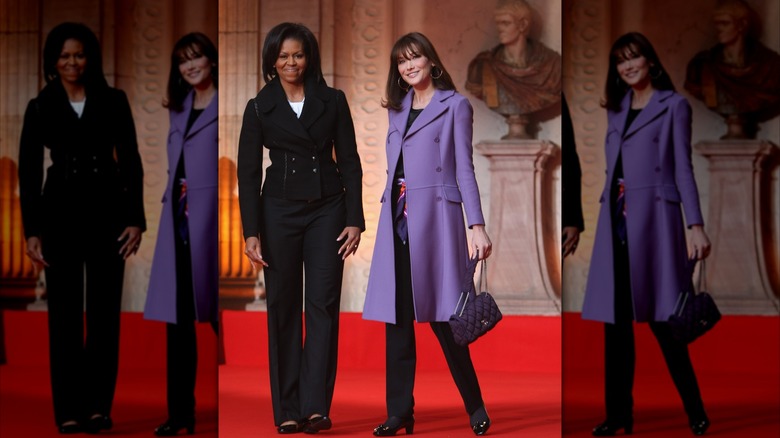 Wearing an all black pants and jacket combo, Michelle Obama smiles while standing next to Carla Bruni-Sarkozy.