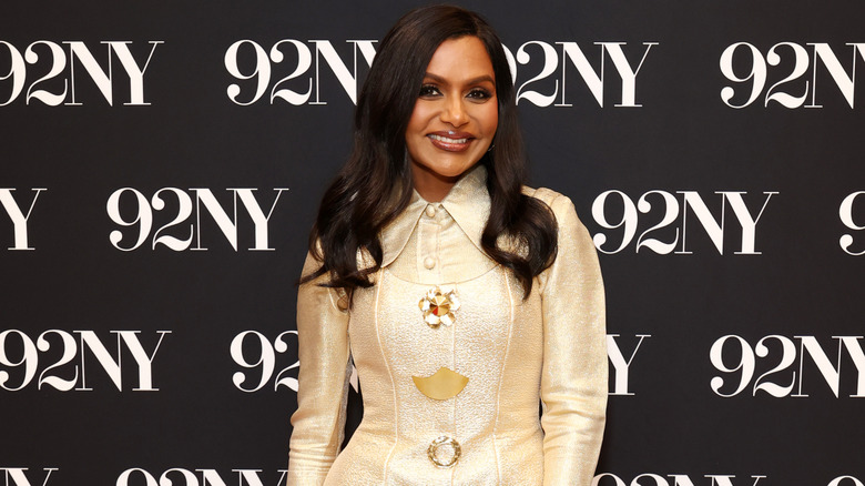 Mindy Kaling smiling at a red carpet event wearing a gold outfit.