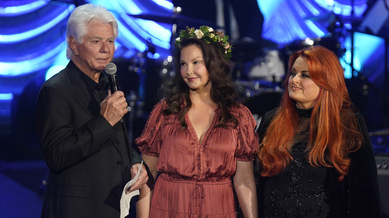 Larry Strickland talking on stage alongside Naomi Judd's daughters