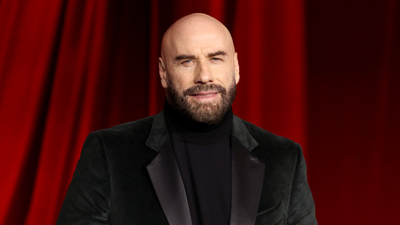 John Travolta smiling and posing in front of red curtain background.