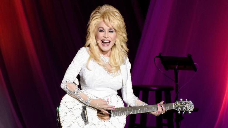 Dolly Parton smiling while performing onstage in an all-white outfit with a white guitar