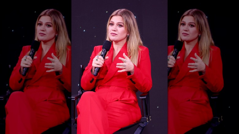 Kelly Clarkson gesturing onstage with a microphone in the other hand in a red suit
