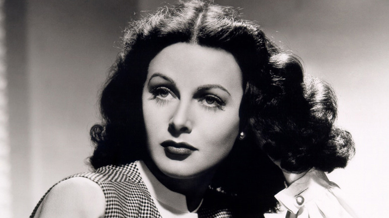Hedy Lamarr, wearing full makeup, looks to the side with one hand touching her hair.