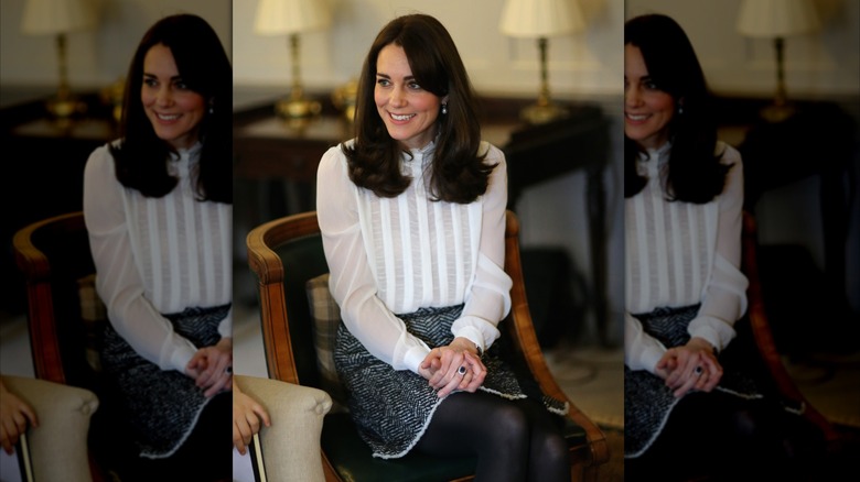Kate Middleton sitting with her legs crossed and wearing a white stripe shirt and herringbone skirt