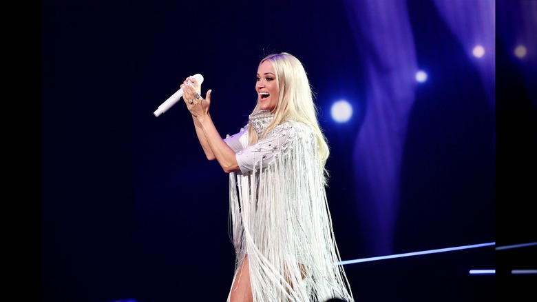 Carrie Underwood claps while wearing a dramatic, fringed outfit.