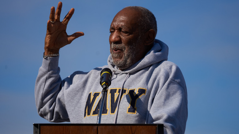 Bill Cosby speaking at a podium in a grey sweatshirt