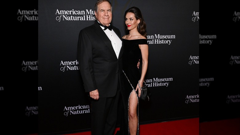 Bill Belichick wearing a tux on the red carpet with Jordon Hudson, who is wearing a black dress with a slit