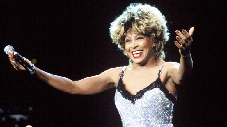 Tina Turner wearing a silver spaghetti-strap dress and smiling with her arms outstretched and microphone in hand during a performance.