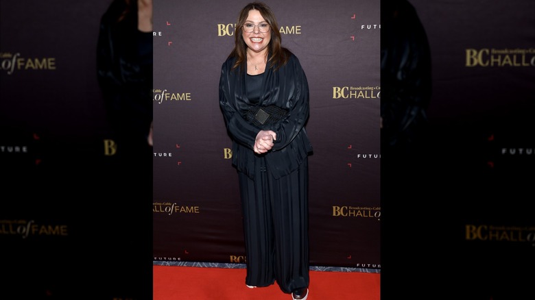 Rachael Ray smiling in a black belted ensemble with glasses on.