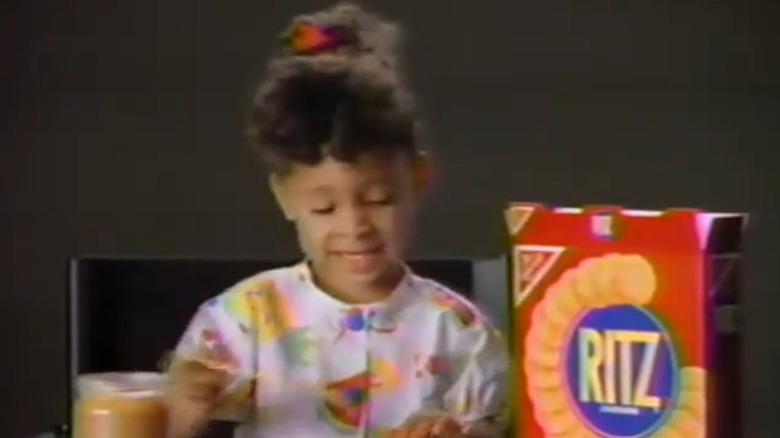 Sporting fuzzy hair and a colorful look, young Raven-Symoné applies peanut butter on a cracker while seated.